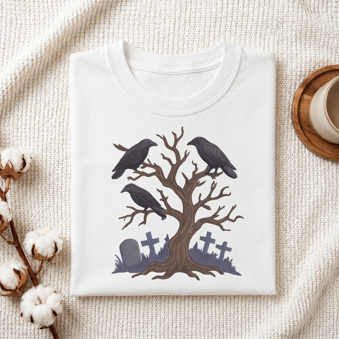 A folded white crewneck t-shirt with a Halloween-themed graphic of two black crows perched on a bare tree with gray tombstones at the base, styled on a textured cream fabric with cotton bolls and a wooden cup.