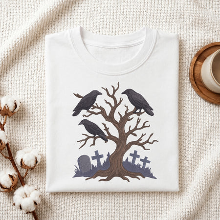 A folded white crewneck t-shirt with a Halloween-themed graphic of two black crows perched on a bare tree with gray tombstones at the base, styled on a textured cream fabric with cotton bolls and a wooden cup.