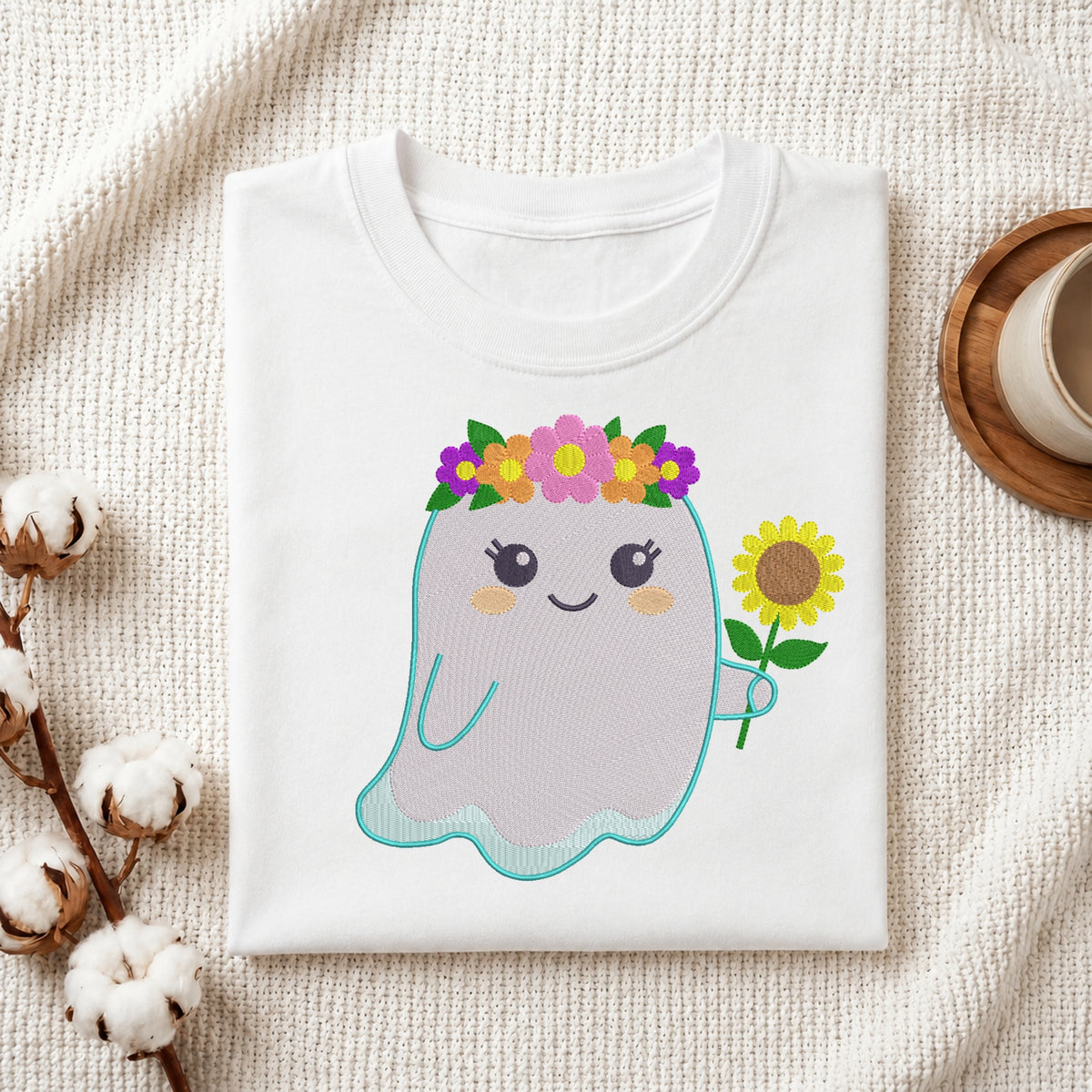 A folded white T-shirt displayed on a textured cream fabric background, featuring a cute embroidered-style ghost graphic with a flower crown and a sunflower. Cotton stems and a small cup on a wooden coaster are placed around it as styled props.