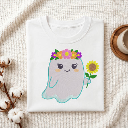 A folded white T-shirt displayed on a textured cream fabric background, featuring a cute embroidered-style ghost graphic with a flower crown and a sunflower. Cotton stems and a small cup on a wooden coaster are placed around it as styled props.