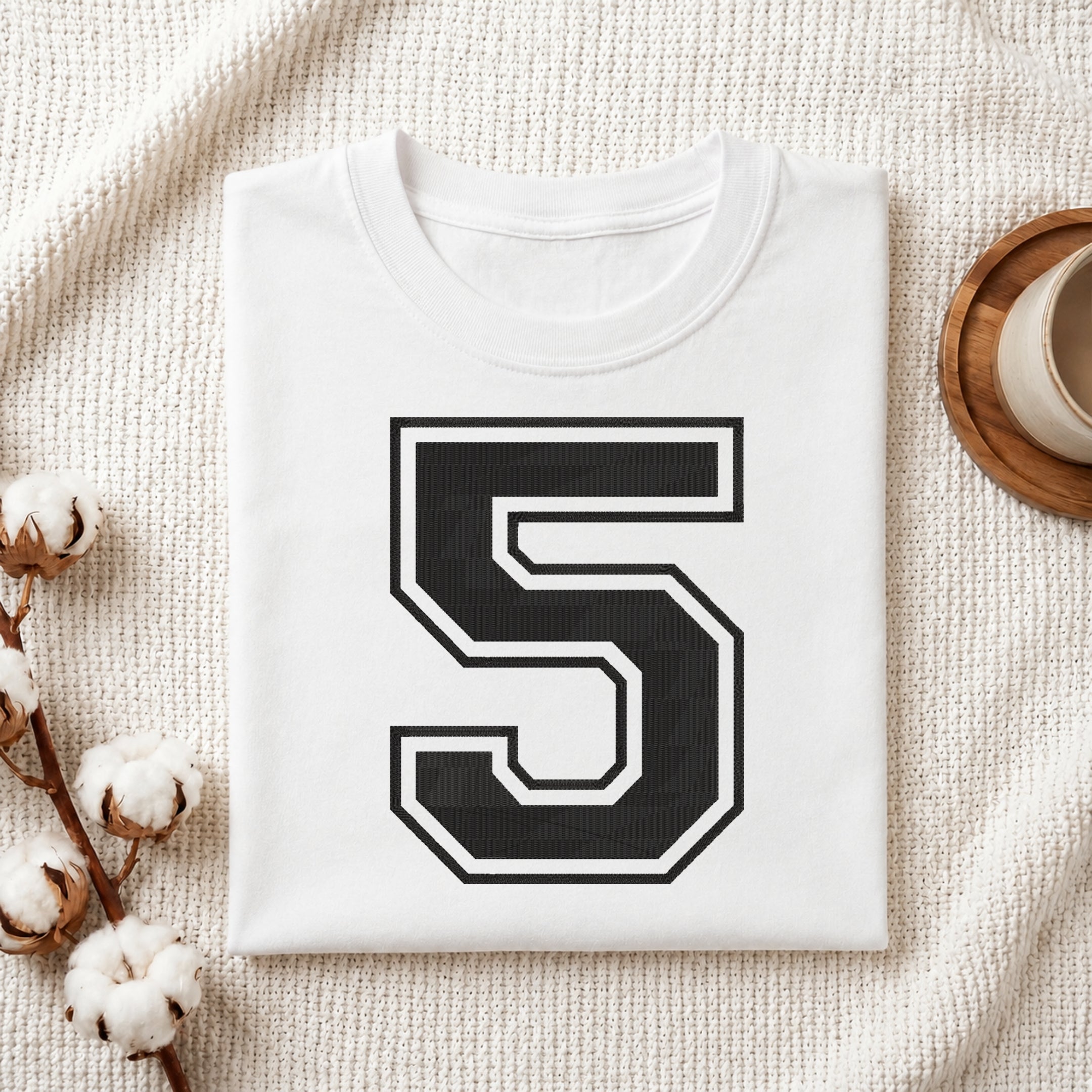 A folded white t-shirt with a large black and white number 5 printed on the front, styled on a textured cream blanket with cotton stems and a cup in the background.
