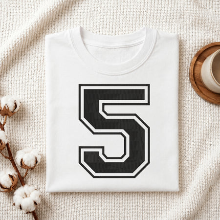 A folded white t-shirt with a large black and white number 5 printed on the front, styled on a textured cream blanket with cotton stems and a cup in the background.