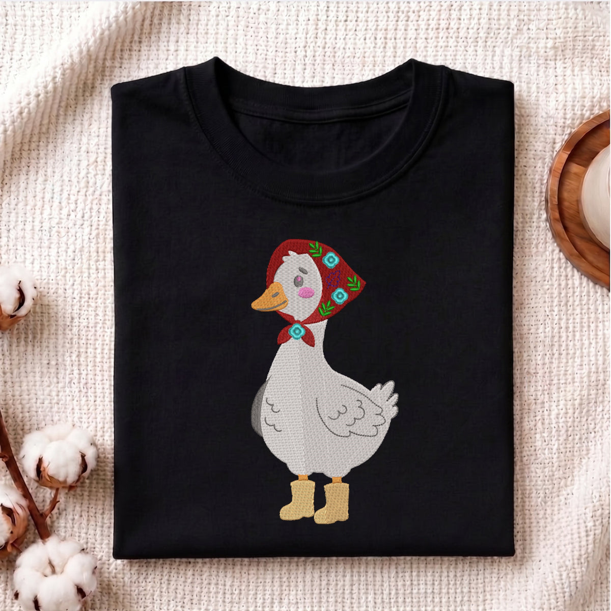 Folded black t-shirt mockup with an embroidery design of a white goose wearing a red headscarf babushka and yellow rainboots.