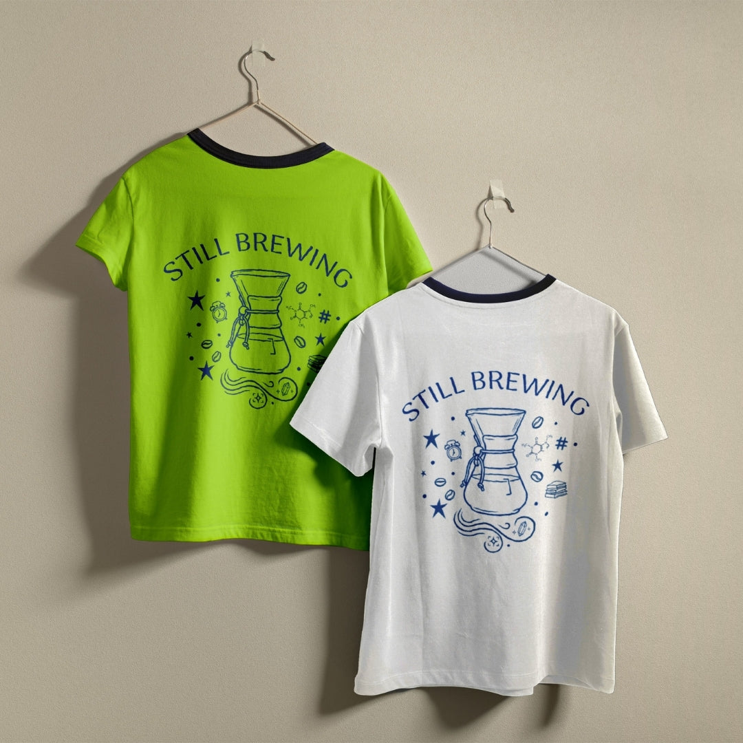 Two t-shirts hanging on a wall, one lime green and one white, both featuring the retro line art "Still Brewing" pour-over carafe graphic design.