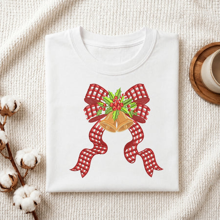 A folded white crewneck t-shirt with a Christmas-themed graphic print featuring a red and white gingham bow, gold bells, and green holly, styled on a textured cream fabric with cotton stems and a coffee cup.