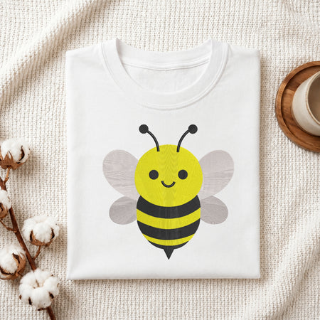 A folded white T-shirt with a cute cartoon bee graphic on the front, styled on a textured cream fabric background with cotton bolls and a small cup in the corners.