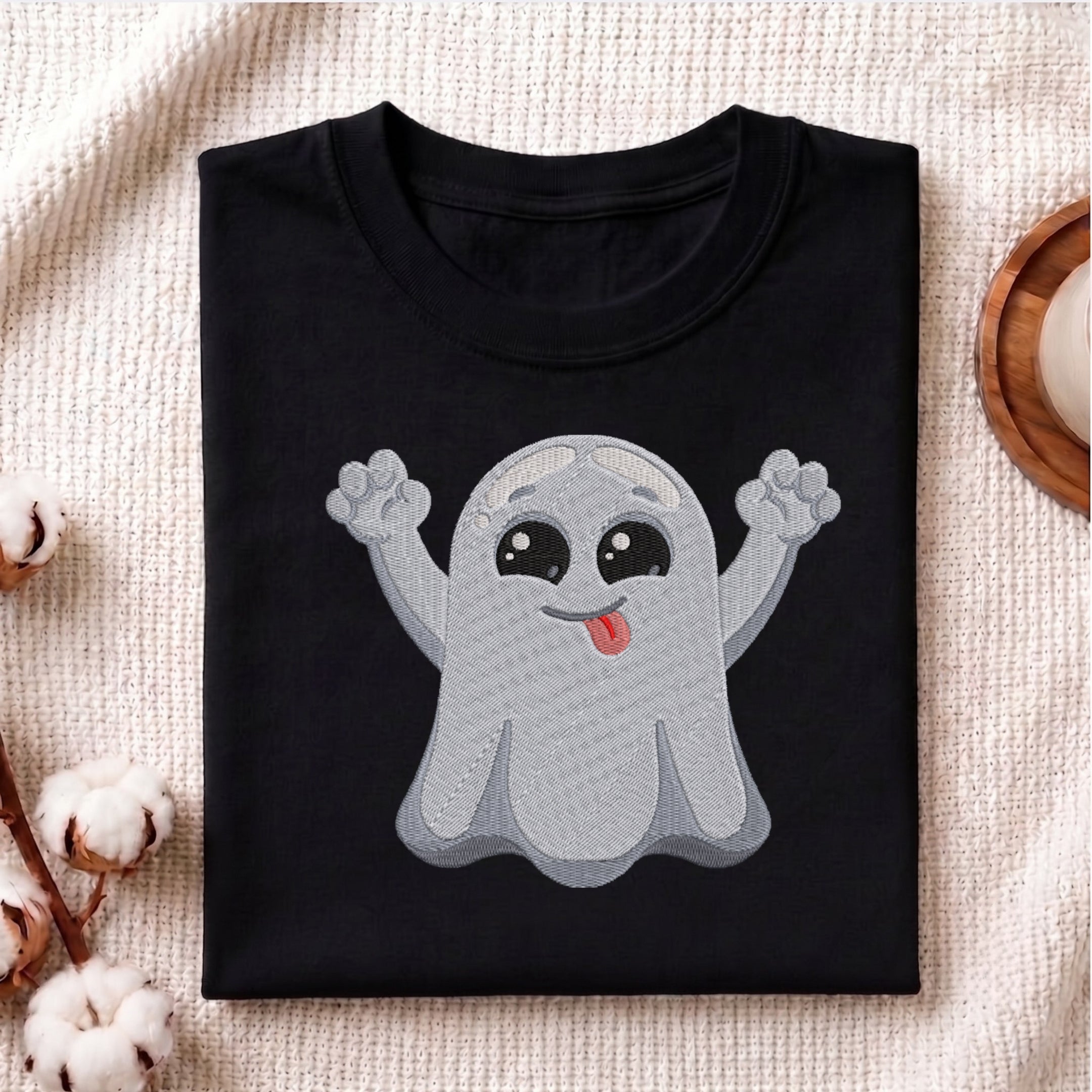A folded black t-shirt with a cute cartoon ghost graphic on the front, displayed on a textured beige fabric background with cotton bolls and a wooden coaster.