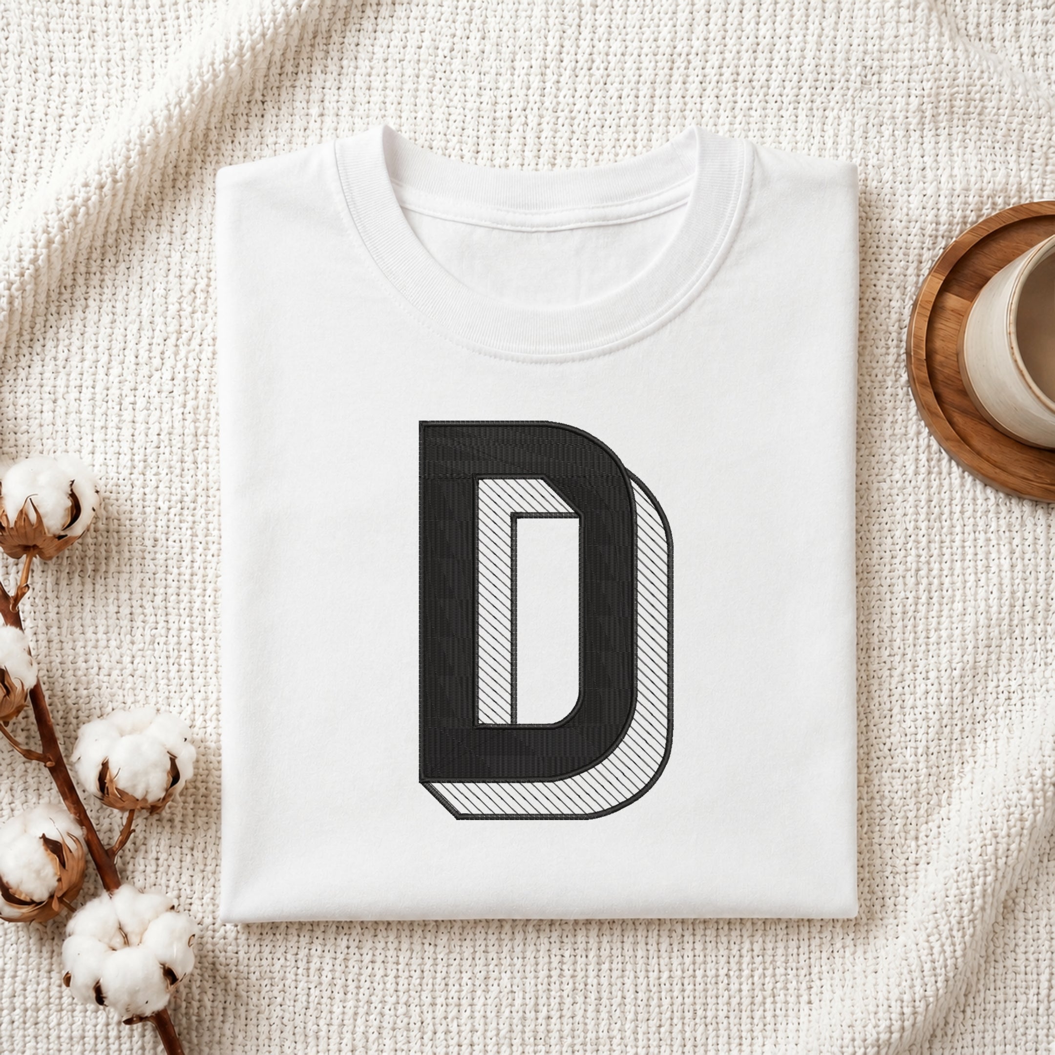 A folded white T-shirt with a large black letter 'D' graphic on the front, styled on a textured cream fabric with cotton bolls and a cup in the background.