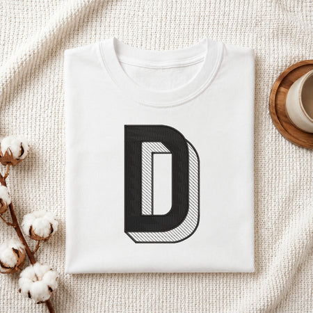 A folded white T-shirt with a large black letter 'D' graphic on the front, styled on a textured cream fabric with cotton bolls and a cup in the background.