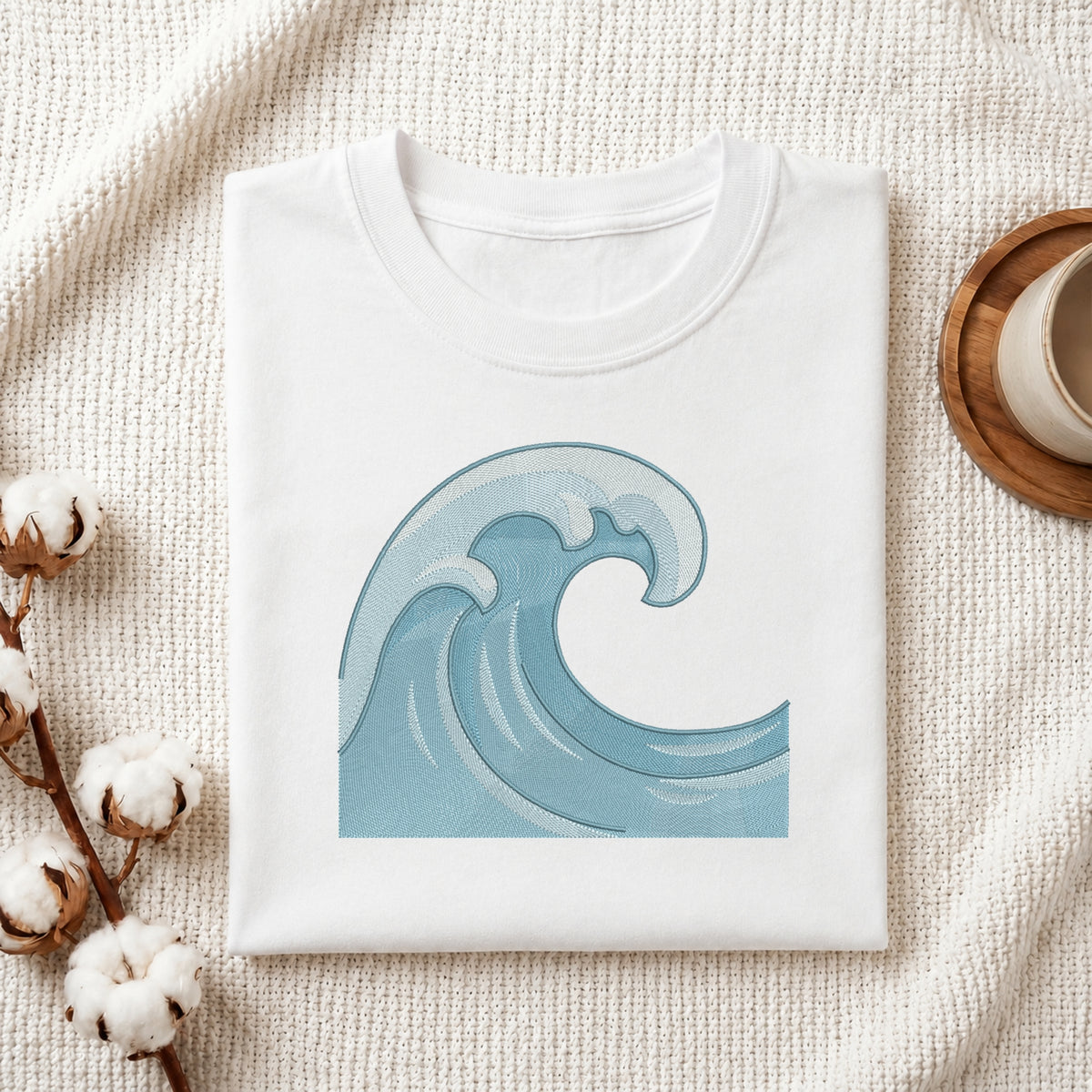 A folded white T-shirt with a light blue ocean wave graphic on the front, styled on a textured cream fabric with cotton bolls and a wooden cup in the background.