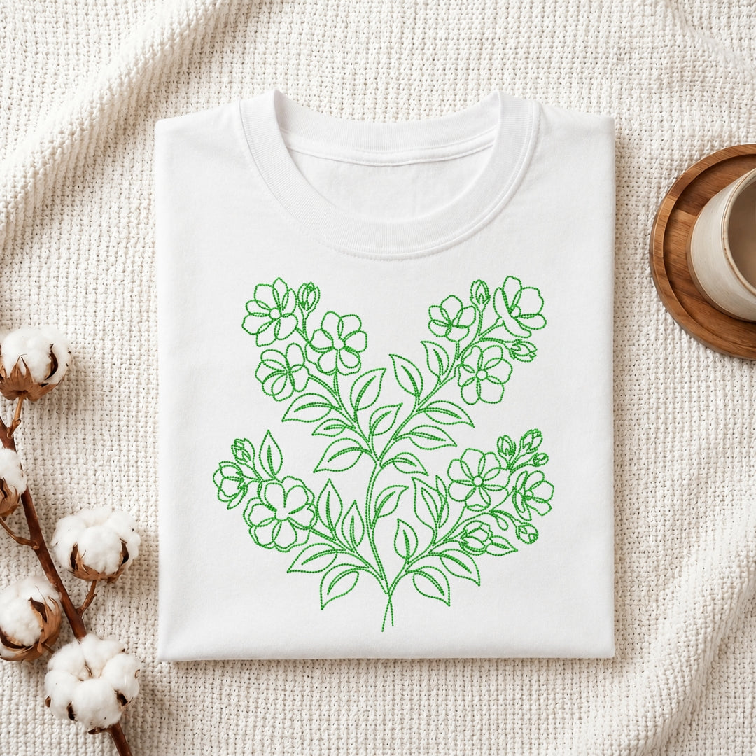 A white t-shirt is folded neatly on a textured white fabric background, featuring a green, line-art floral embroidery design in the center.
