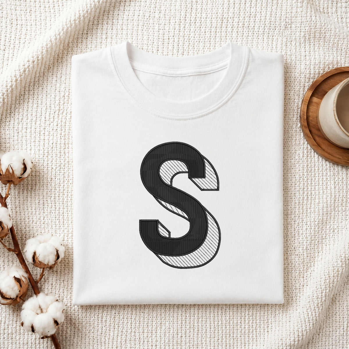 A folded white T-shirt with a large black decorative letter 'S' printed on the front, styled on a textured cream fabric with cotton stems and a small cup in the corners.