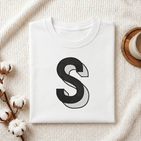 A folded white T-shirt with a large black decorative letter 'S' printed on the front, styled on a textured cream fabric with cotton stems and a small cup in the corners.