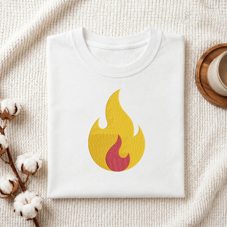 A folded white t-shirt with a yellow and red flame graphic printed on the chest, styled on a textured cream fabric with cotton bolls and a wooden cup in the background.