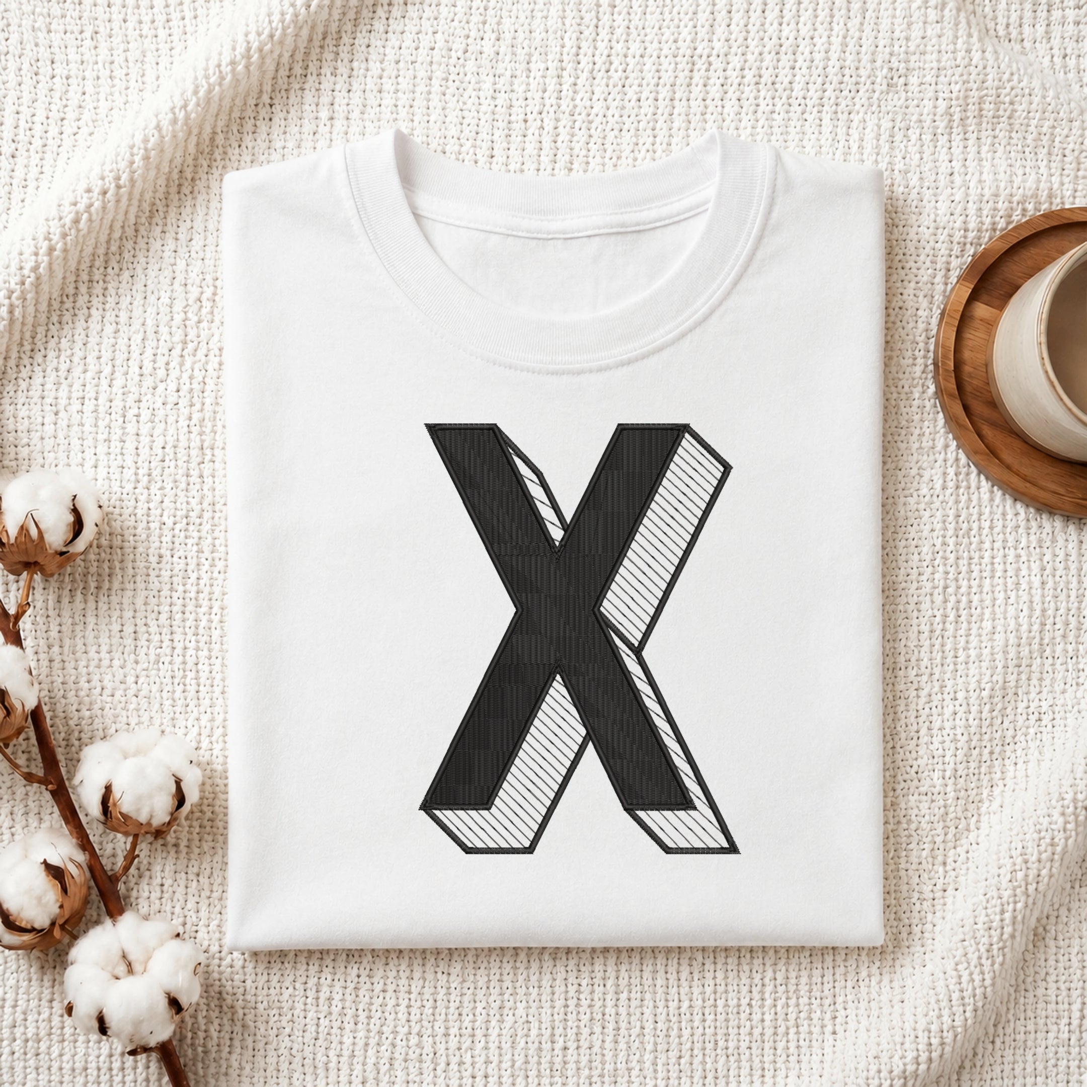 A neatly folded white crewneck t-shirt featuring a stitched 3D block letter "X" design, styled with cotton branches and a wooden coaster.