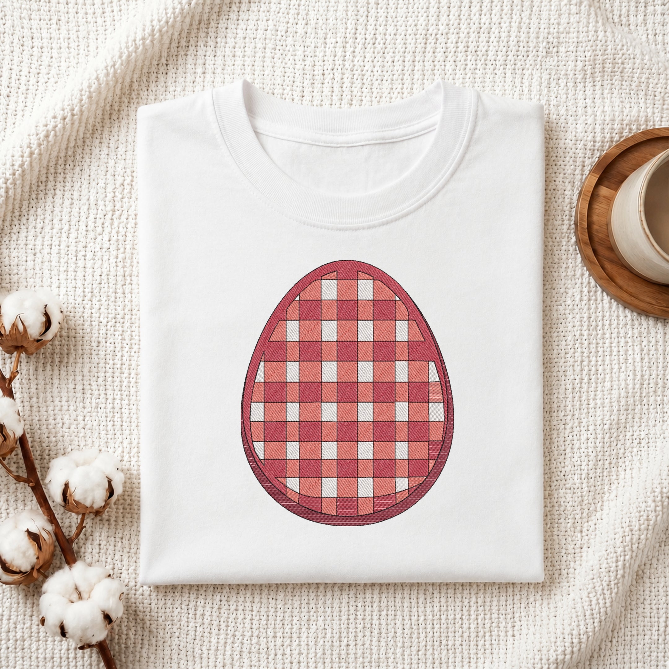 A folded white T-shirt laid flat on a textured cream fabric background, featuring a large red and pink gingham check egg graphic on the chest. Cotton bolls and a small cup on a wooden coaster are placed around it as styled props.