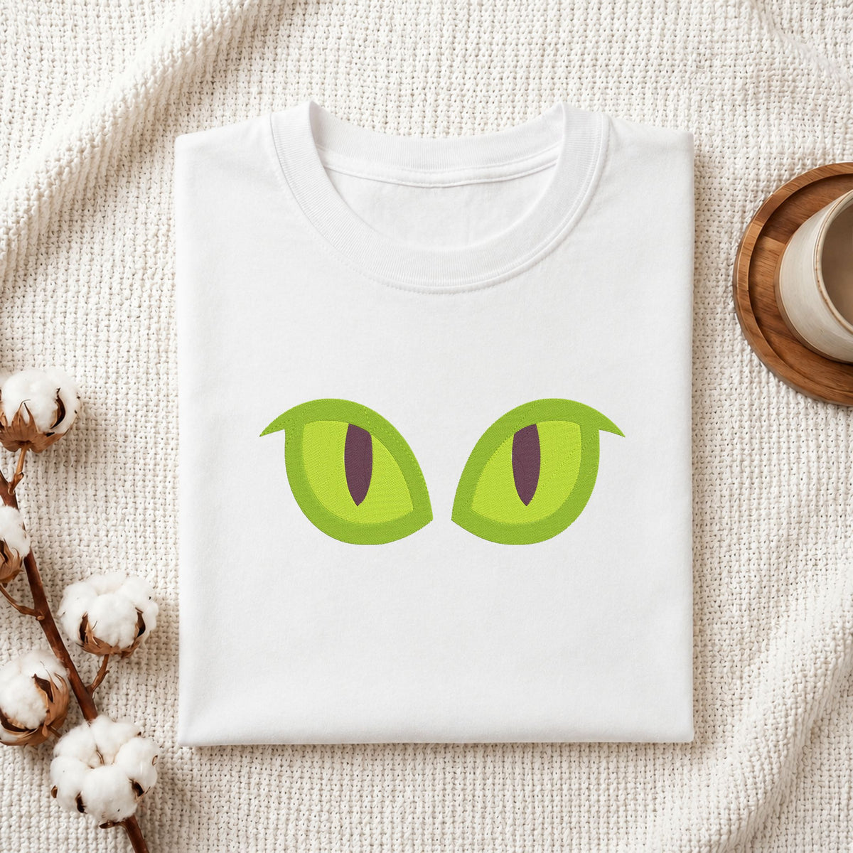 A folded white t-shirt with a graphic of two green cat eyes, styled on a textured cream knit blanket with cotton bolls and a cup in the background.