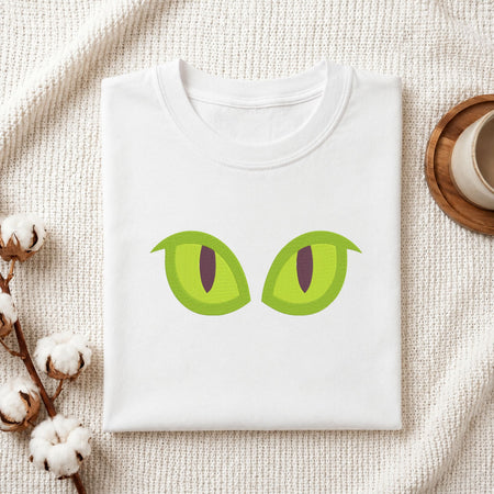 A folded white t-shirt with a graphic of two green cat eyes, styled on a textured cream knit blanket with cotton bolls and a cup in the background.
