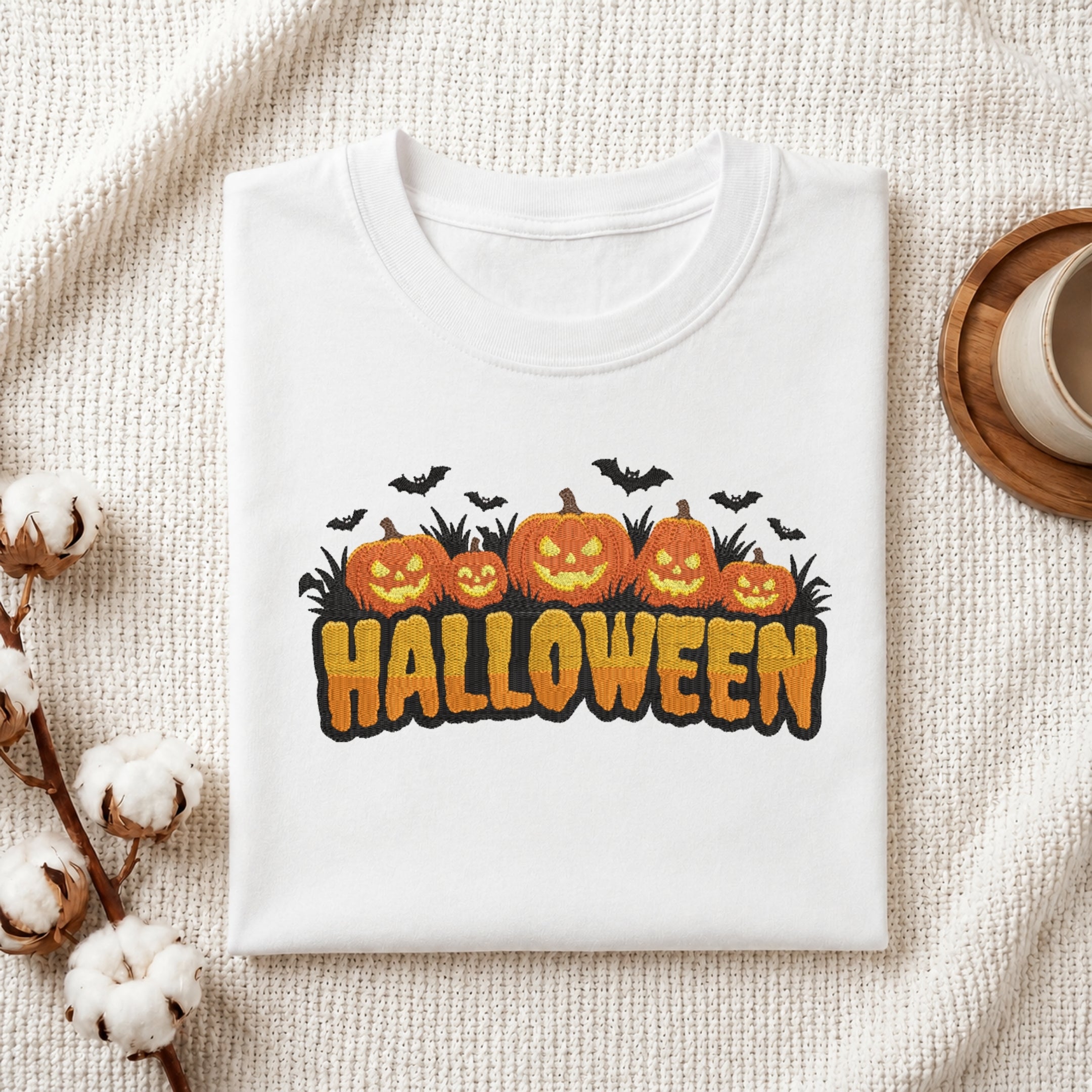 A folded white t-shirt with a Halloween graphic print featuring jack-o'-lanterns, bats, and the word 'HALLOWEEN' in orange, displayed on a textured cream fabric with cotton stems and a cup in the background.
