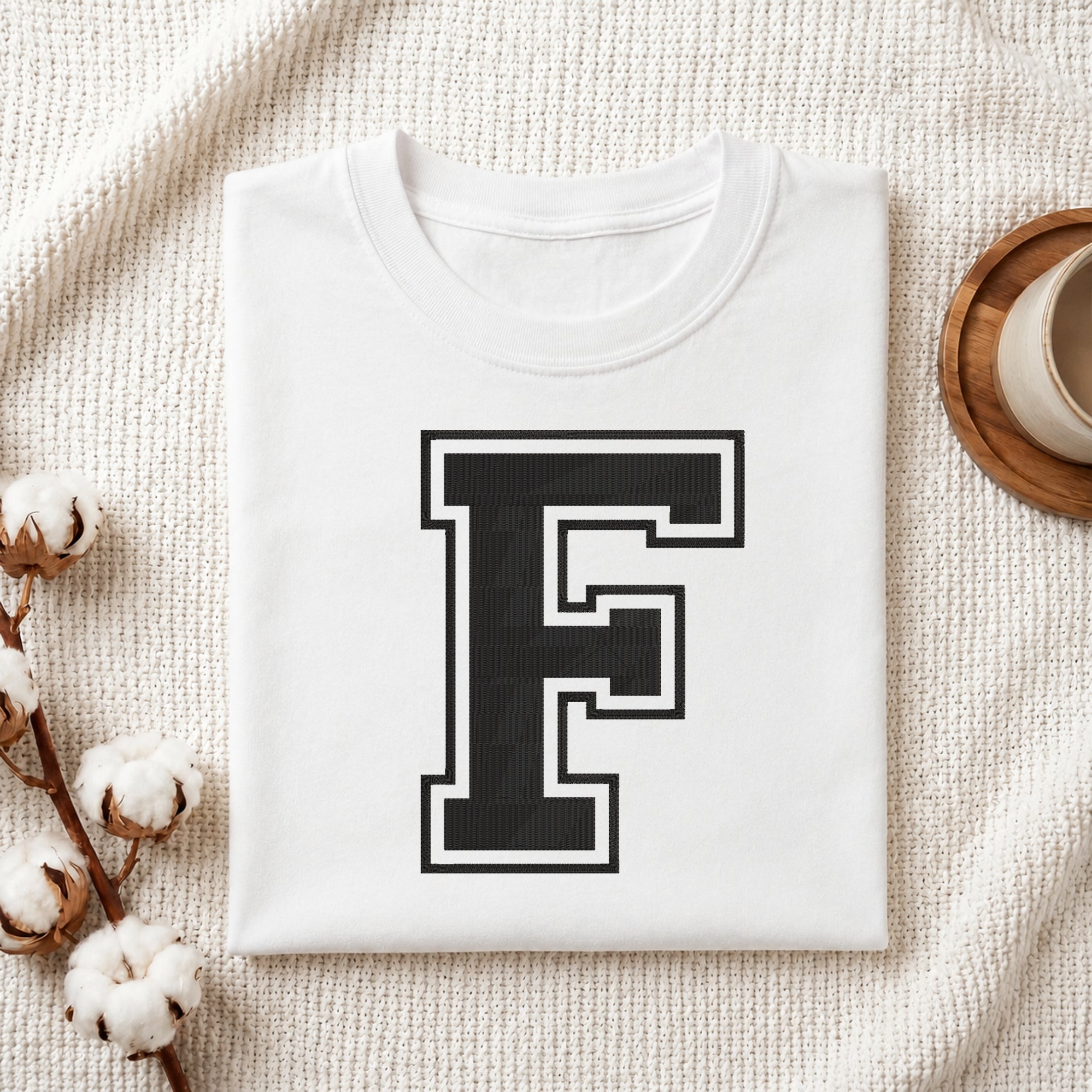 A folded white T-shirt with a large black block letter 'F' outlined in white on the front, styled on a textured cream fabric with cotton bolls and a small cup in the background.