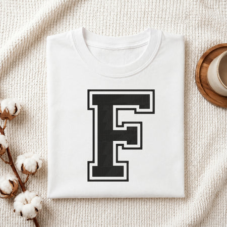 A folded white T-shirt with a large black block letter 'F' outlined in white on the front, styled on a textured cream fabric with cotton bolls and a small cup in the background.