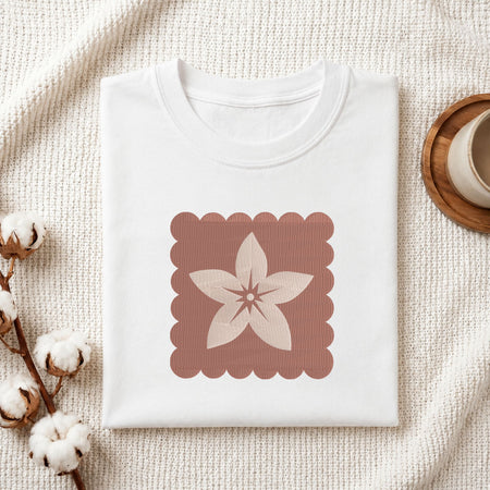 Folded white cotton t-shirt mockup featuring a minimalist flower machine embroidery design, styled with cotton stems and a wooden cup.