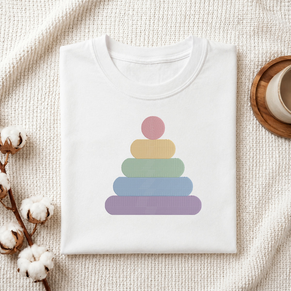 A folded white t-shirt with a pastel rainbow stacking graphic, styled on a textured cream fabric with cotton bolls and a wooden cup in the background.
