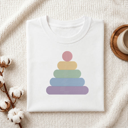 A folded white t-shirt with a pastel rainbow stacking graphic, styled on a textured cream fabric with cotton bolls and a wooden cup in the background.
