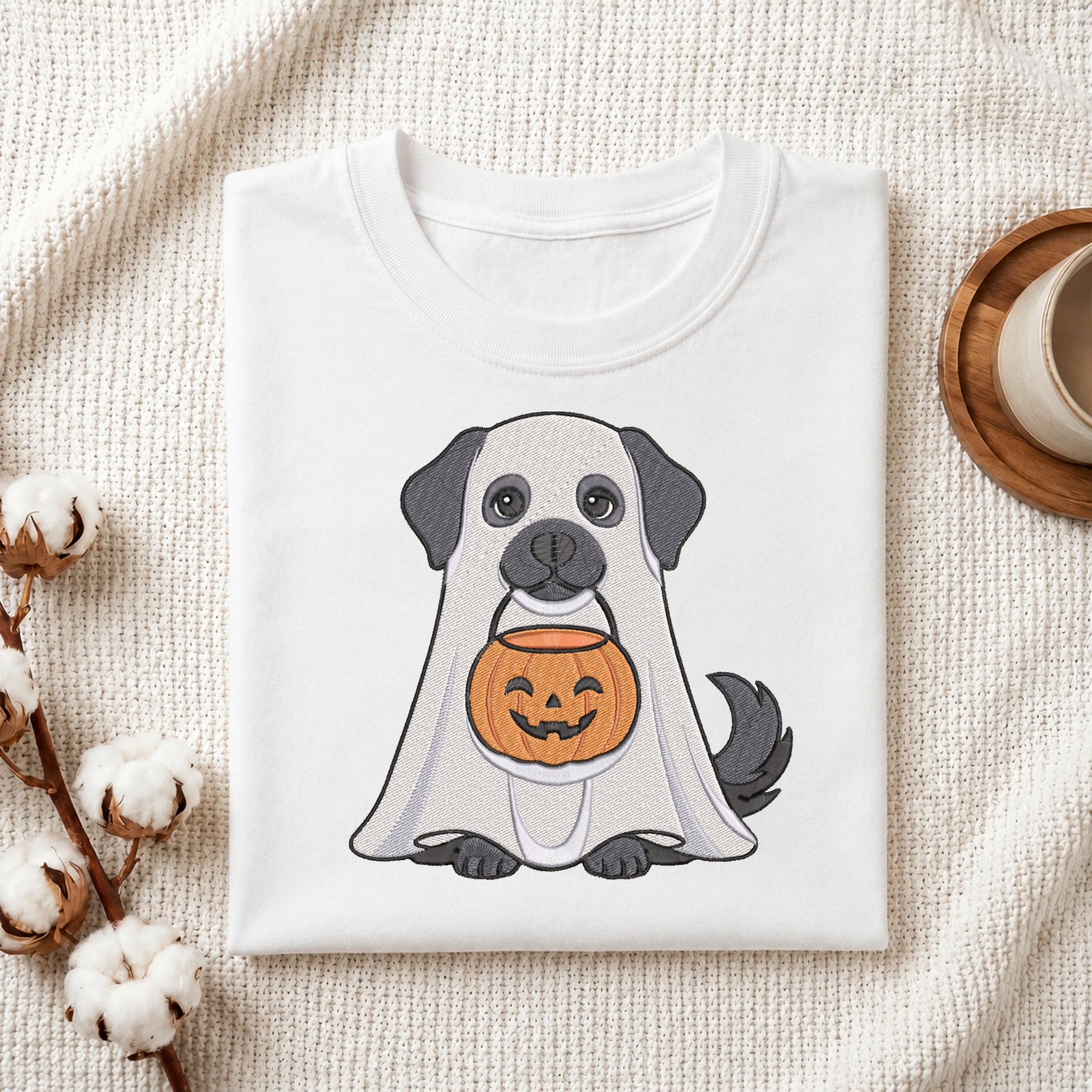 A folded white T-shirt with a cute cartoon dog dressed as a ghost and holding a jack-o'-lantern, styled on a textured cream fabric with cotton stems and a cup in the background.