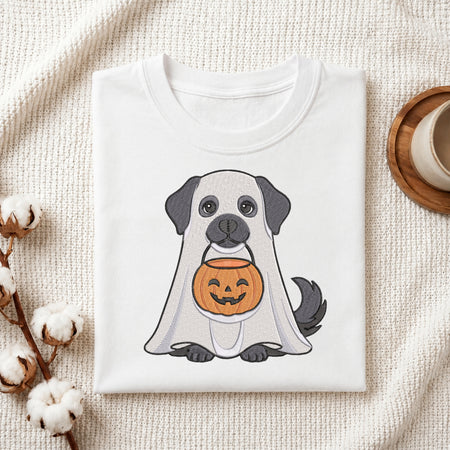 A folded white T-shirt with a cute cartoon dog dressed as a ghost and holding a jack-o'-lantern, styled on a textured cream fabric with cotton stems and a cup in the background.