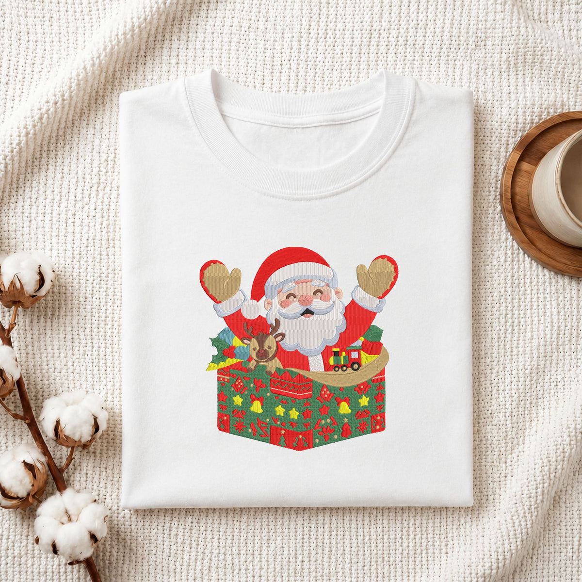 A folded white crewneck t-shirt with a colorful Christmas graphic of Santa Claus popping out of a gift box, styled on a textured cream fabric with cotton stems and a coffee cup in the background.
