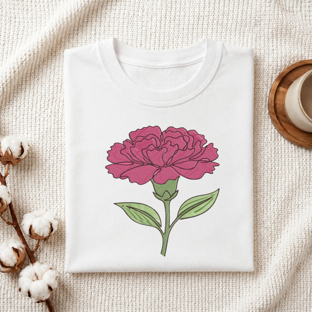 A folded white crewneck t-shirt featuring a graphic of a pink carnation flower with green leaves, styled on a textured cream fabric with cotton bolls and a wooden cup.