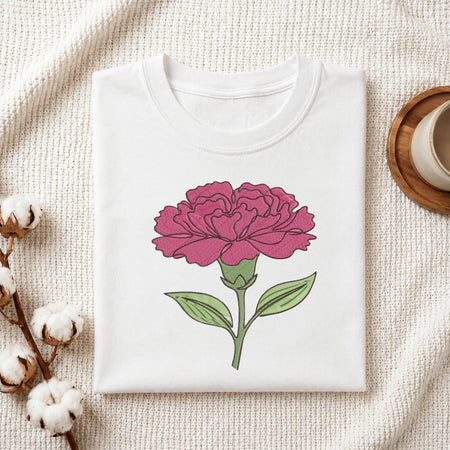 A folded white crewneck t-shirt featuring a graphic of a pink carnation flower with green leaves, styled on a textured cream fabric with cotton bolls and a wooden cup.