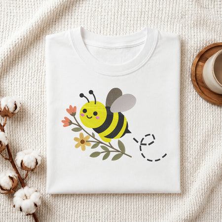 A folded white t-shirt with a cute cartoon bee and flower graphic, styled on a textured cream fabric with cotton bolls and a wooden cup in the background.