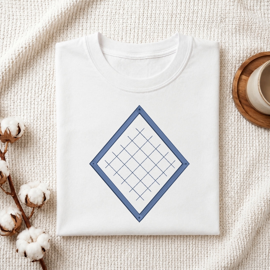 A folded white crewneck t-shirt with a blue diamond grid graphic on the front, styled on a textured cream knit blanket with cotton bolls and a wooden cup in the background.