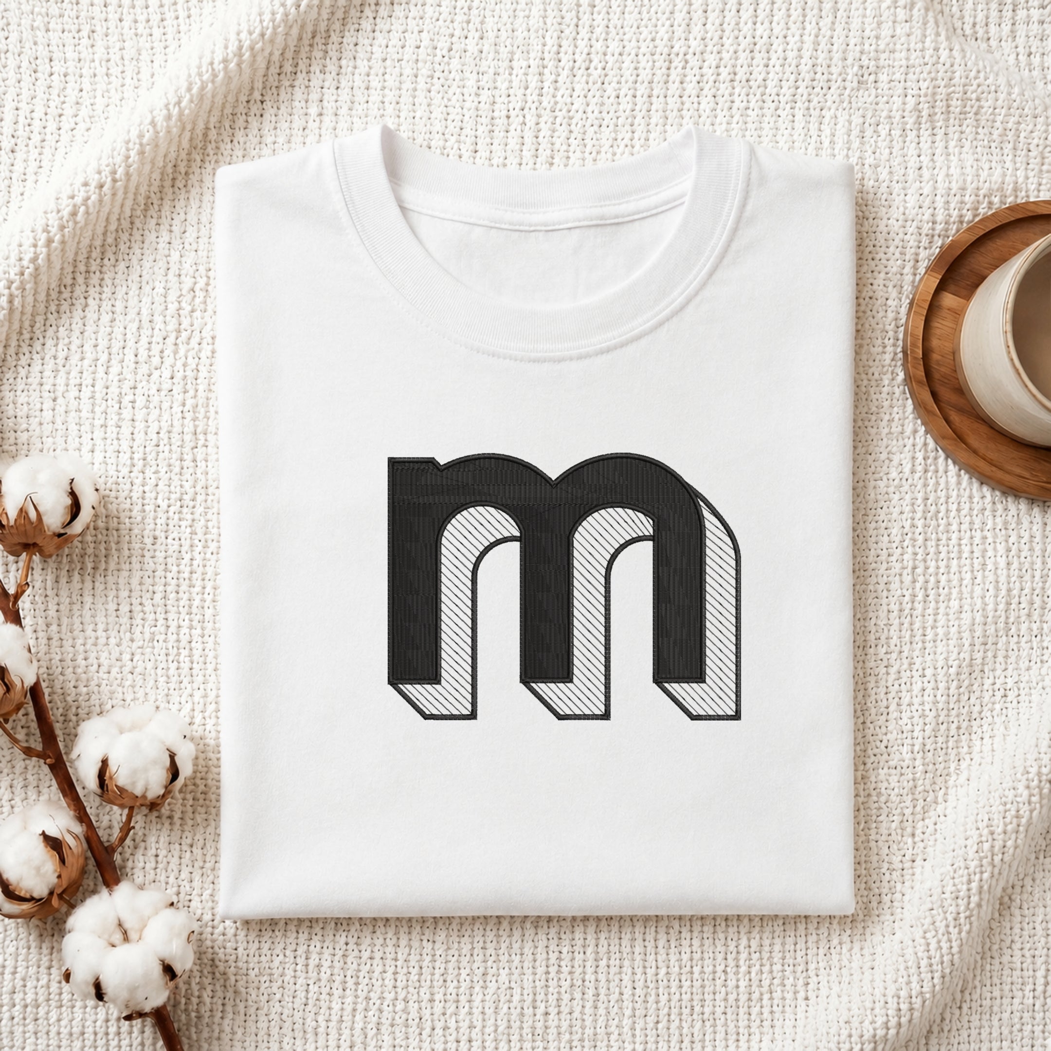 A folded white t-shirt with a large black stylized letter 'm' graphic on the front, displayed on a textured cream knit blanket with cotton bolls and a cup of coffee in the background.