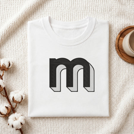 A folded white t-shirt with a large black stylized letter 'm' graphic on the front, displayed on a textured cream knit blanket with cotton bolls and a cup of coffee in the background.