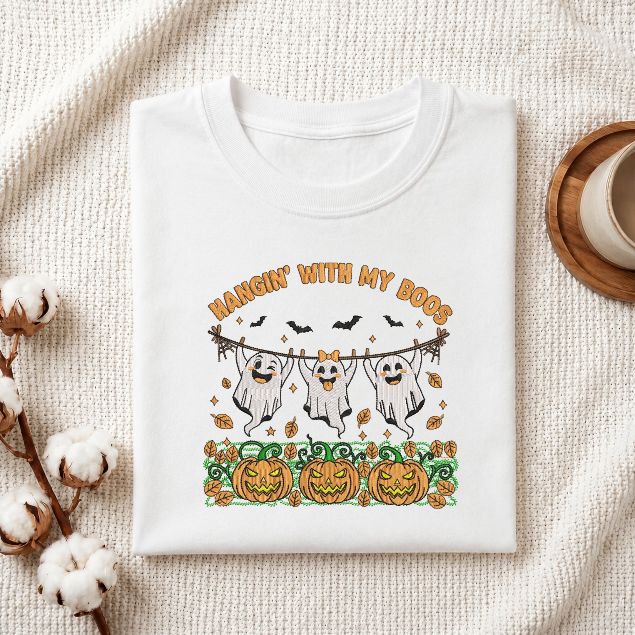 A folded white graphic T-shirt displayed on a textured cream fabric background with styled props, including cotton stems and a wooden cup. The shirt features a Halloween-themed print with the text 'HANGIN' WITH MY DOGS', three cartoon ghosts hanging from a rope, bats, pumpkins, and autumn leaves.