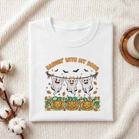 A folded white graphic T-shirt displayed on a textured cream fabric background with styled props, including cotton stems and a wooden cup. The shirt features a Halloween-themed print with the text 'HANGIN' WITH MY DOGS', three cartoon ghosts hanging from a rope, bats, pumpkins, and autumn leaves.