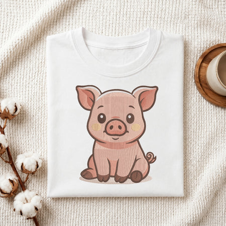 A folded white crewneck t-shirt featuring a cute cartoon illustration of a pink pig, styled on a textured cream fabric with cotton bolls and a wooden cup in the background.