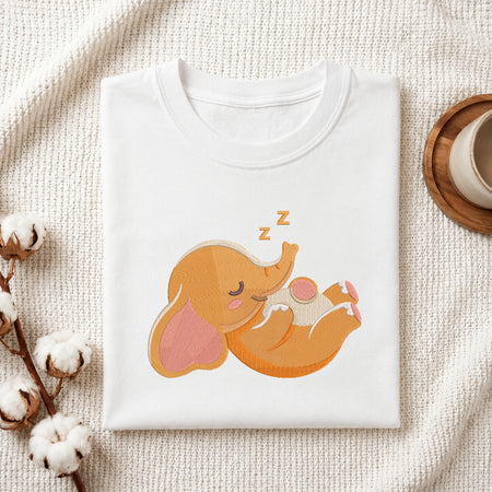 A folded white crewneck t-shirt featuring a cute cartoon sleeping elephant graphic in orange, pink, and beige, styled on a textured cream fabric with cotton bolls and a wooden cup.