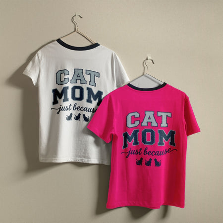 Two t-shirts hanging on a wall, one white and one hot pink, both featuring the retro collegiate "Cat Mom just because" design with cat silhouettes.
