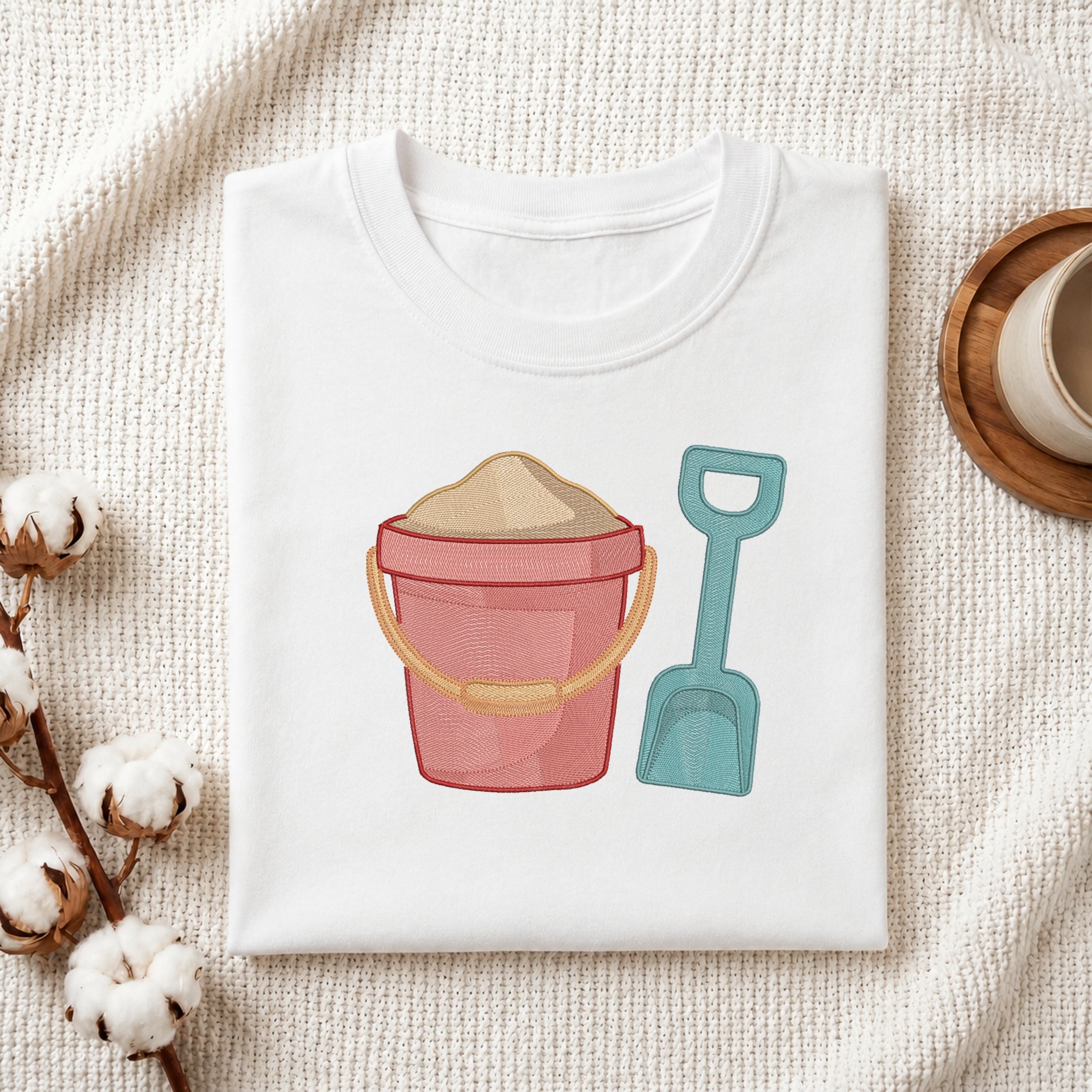 A folded white T-shirt with a beach-themed graphic of a pink bucket filled with sand and a teal shovel, styled on a textured cream fabric with cotton bolls and a wooden cup.