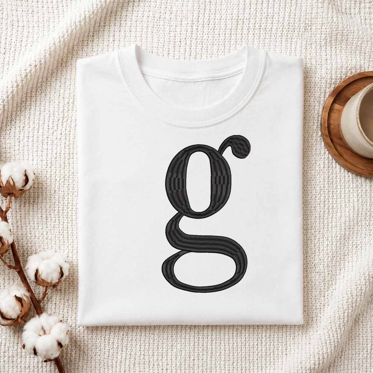 A folded white crewneck t-shirt with a large black stylized letter 'g' printed on the front, styled on a textured cream knit blanket with cotton bolls and a wooden cup in the background.
