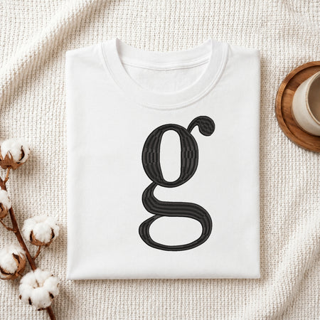 A folded white crewneck t-shirt with a large black stylized letter 'g' printed on the front, styled on a textured cream knit blanket with cotton bolls and a wooden cup in the background.
