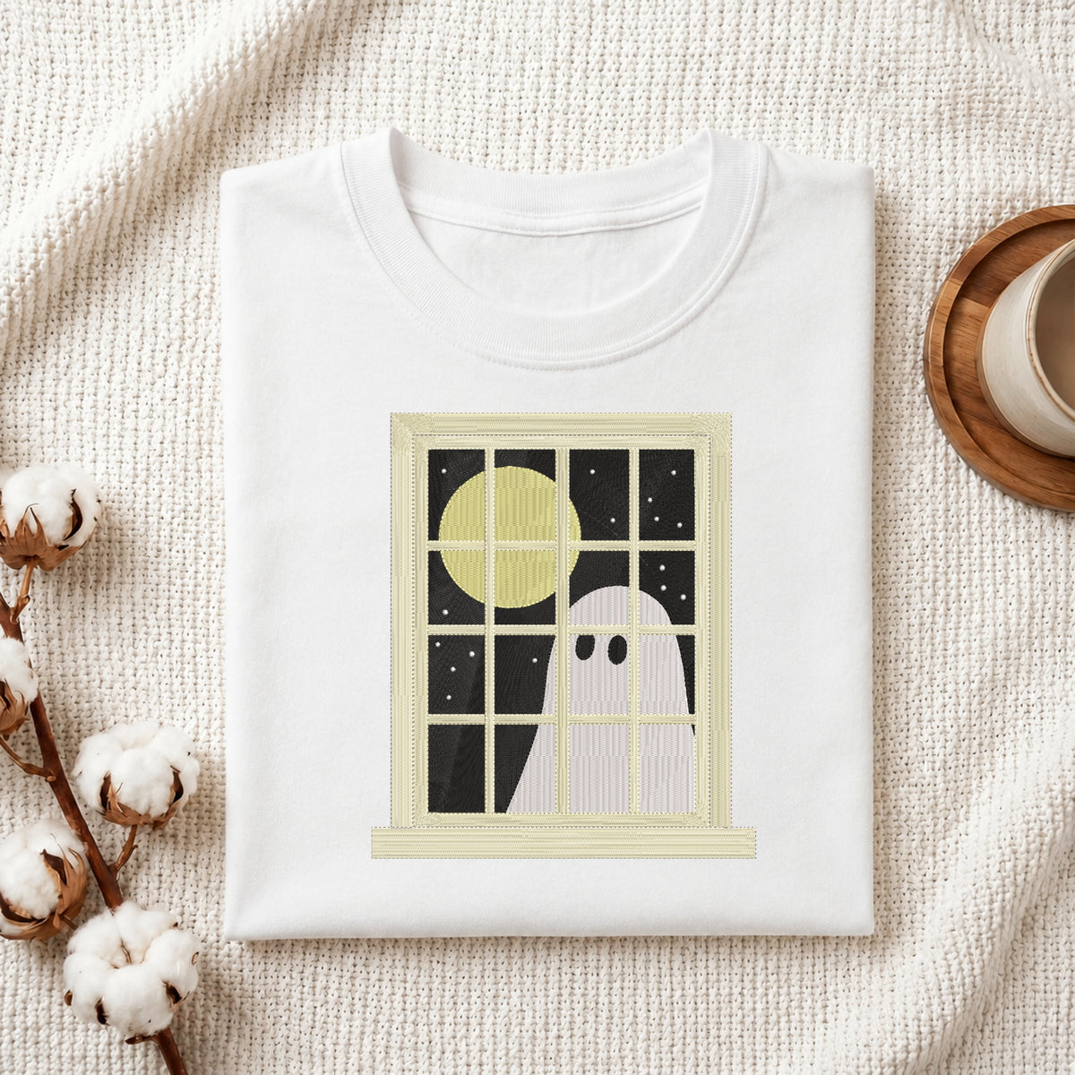 A folded white crewneck t-shirt with a graphic print of a ghost looking out a window at a full moon, styled on a textured cream knit blanket with cotton bolls and a coffee cup.