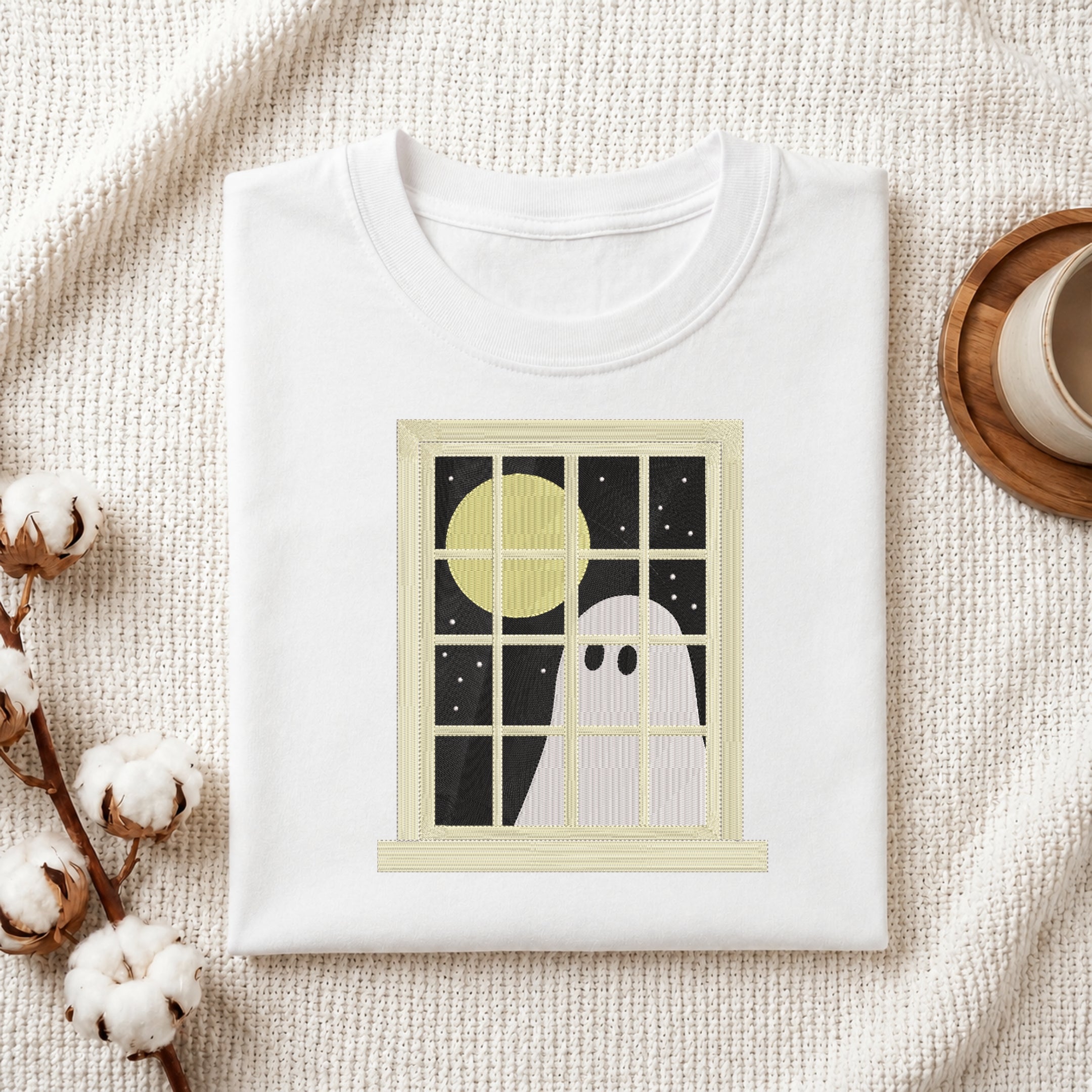 A folded white crewneck t-shirt with a graphic print of a ghost looking out a window at a full moon, styled on a textured cream knit blanket with cotton bolls and a coffee cup.