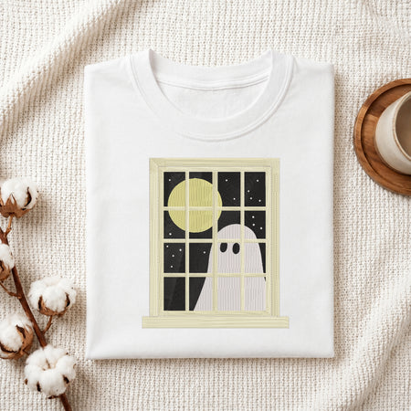 A folded white crewneck t-shirt with a graphic print of a ghost looking out a window at a full moon, styled on a textured cream knit blanket with cotton bolls and a coffee cup.