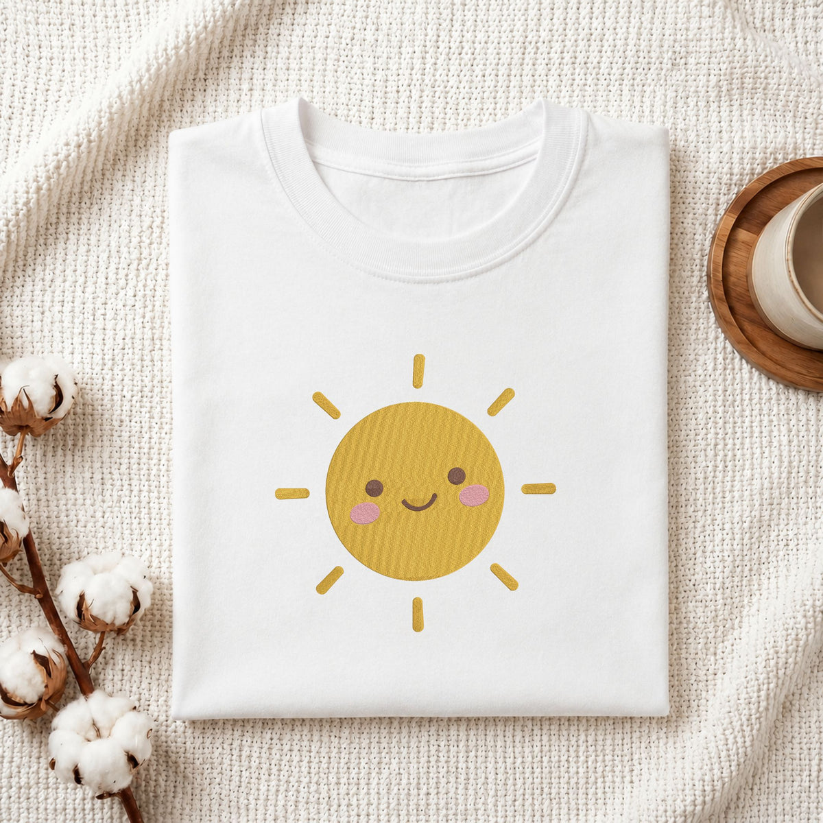 Folded white cotton t-shirt mockup featuring a cute smiling sun machine embroidery design, styled with cotton stems and a wooden cup   