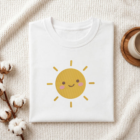 Folded white cotton t-shirt mockup featuring a cute smiling sun machine embroidery design, styled with cotton stems and a wooden cup   